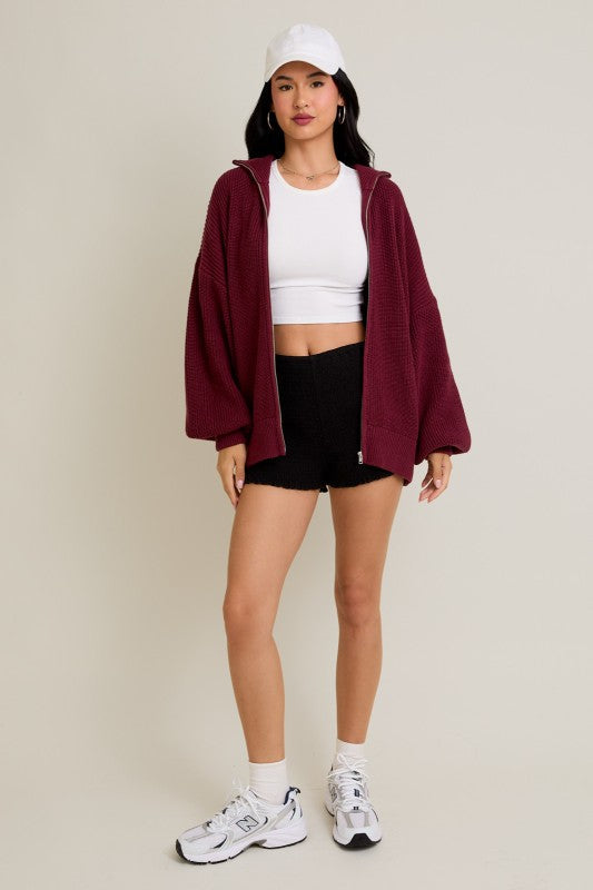 Person wearing a burgundy jacket, white crop top, black shorts, and white sneakers on a beige background