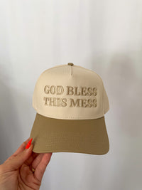 Beige cap with 'God Bless This Mess' text held by a hand against a neutral background