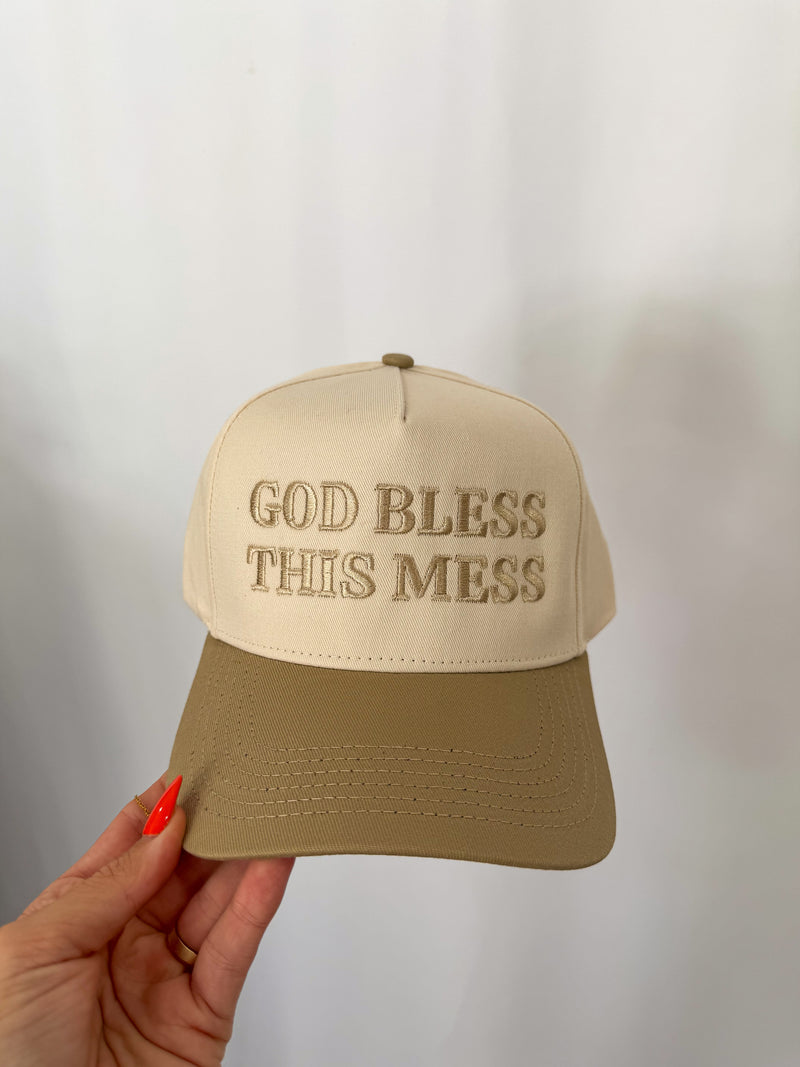 Beige cap with 'God Bless This Mess' text held by a hand against a neutral background