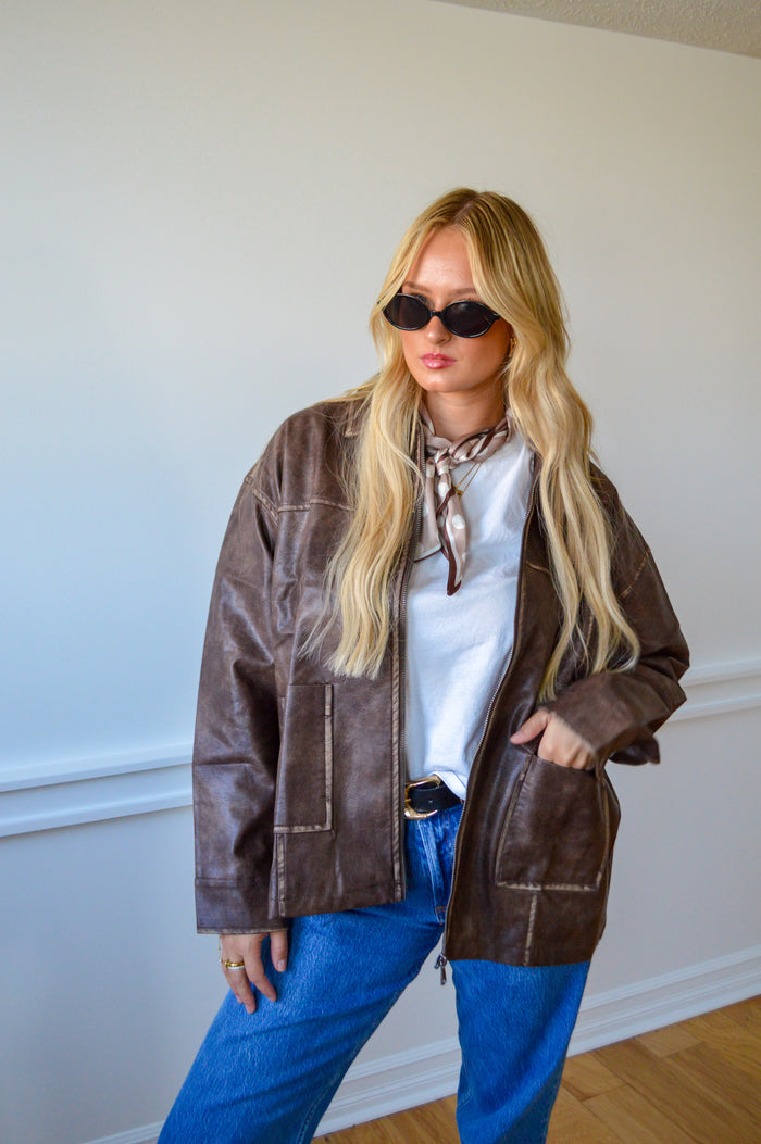 Woman wearing a brown leather jacket, white shirt, and blue jeans against a plain wall.