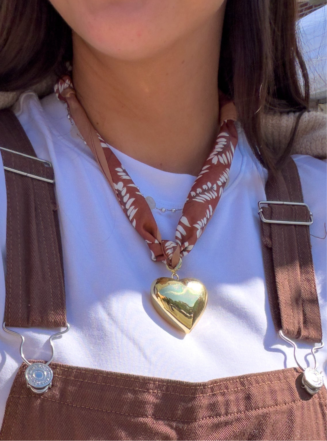 Person wearing a gold heart-shaped pendant necklace with a white shirt and brown overalls.