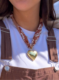 Person wearing a gold heart-shaped pendant necklace with a white shirt and brown overalls.
